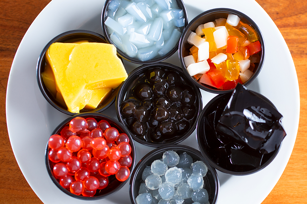 Assorted Toppings for Bubble Tea and Boba Tea: “Variety of toppings for bubble tea and boba tea, including fruit jelly, tapioca pearls, popping boba, and grass jelly, displayed in small bowls on a plate.”
