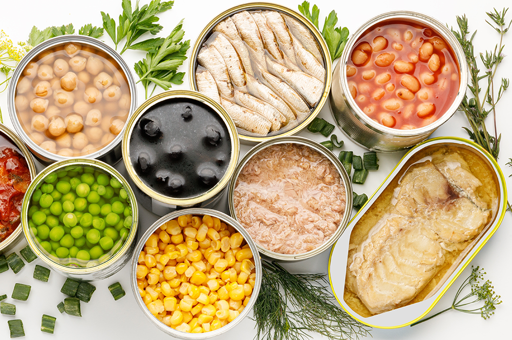 Various canned convenience foods including beans, corn, peas, tuna, and tomatoes along with fresh herbs, offering quick meal options.
