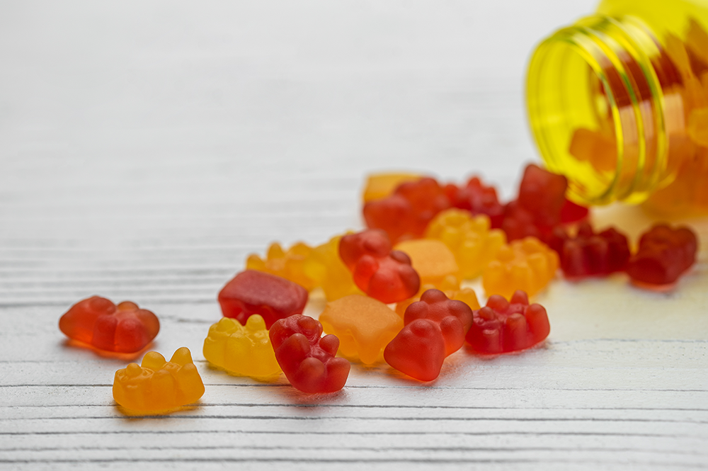 A close-up view of gummy bear candies in vibrant shades of red, orange, and yellow, spilling out of a clear pill bottle onto a white wooden surface, emphasizing the bright, eye-catching colors of these popular gummy treats. Gummies from Symega