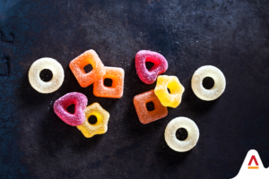 A scattered arrangement of sugar-coated gummy rings in pink, orange, and yellow colors, contrasting against a dark, textured background and interspersed with plain white gummy rings, creating an visually striking composition of vibrant gummy candies. Gummies from Symega